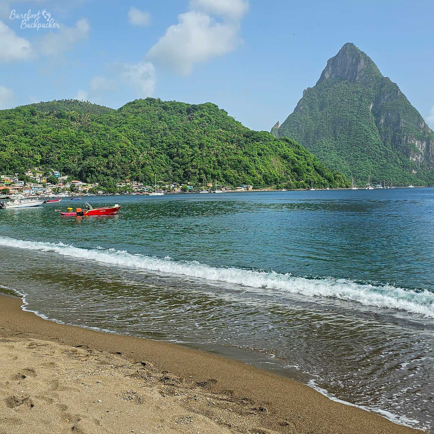 View of a beach and ocean with a large mountain in the background, covered in trees. A red boat floats on the water.