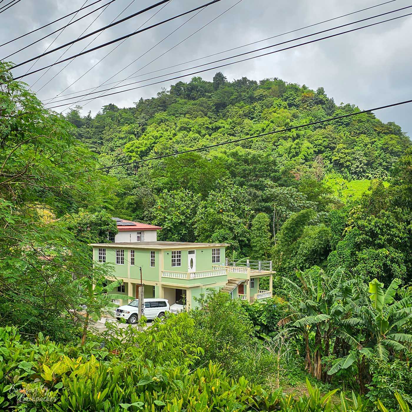 A light-green two-story house with a white balcony and white vehicle parked outside, set against a lush, green, forested hillside beneath a cloudy sky, seen from a slightly obscured angle through dense foliage.