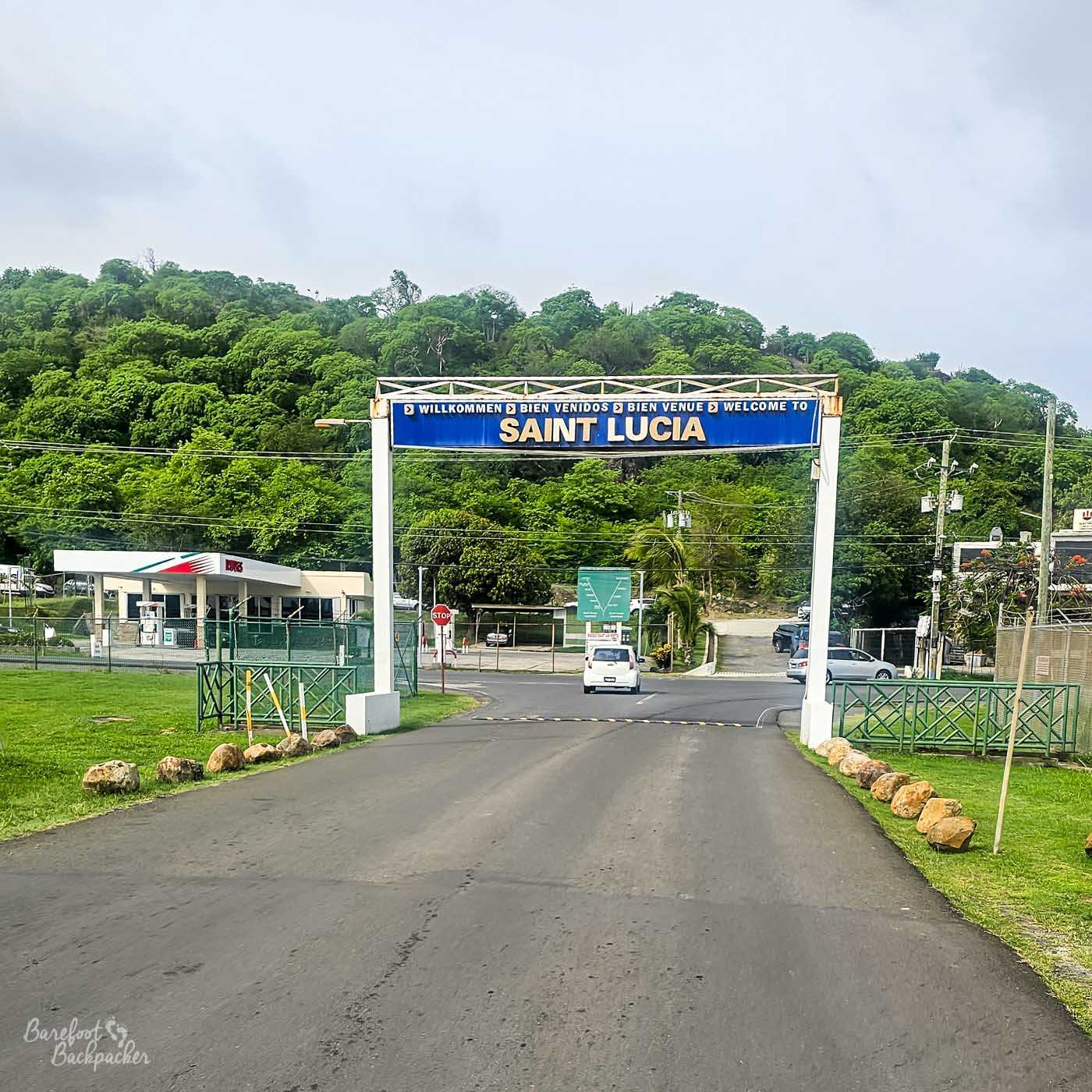 A large roadside archway sign reading “Welcome to Saint Lucia” in multiple languages spans a paved road. Cars pass beneath it, with lush green hills and roadside buildings visible in the background.