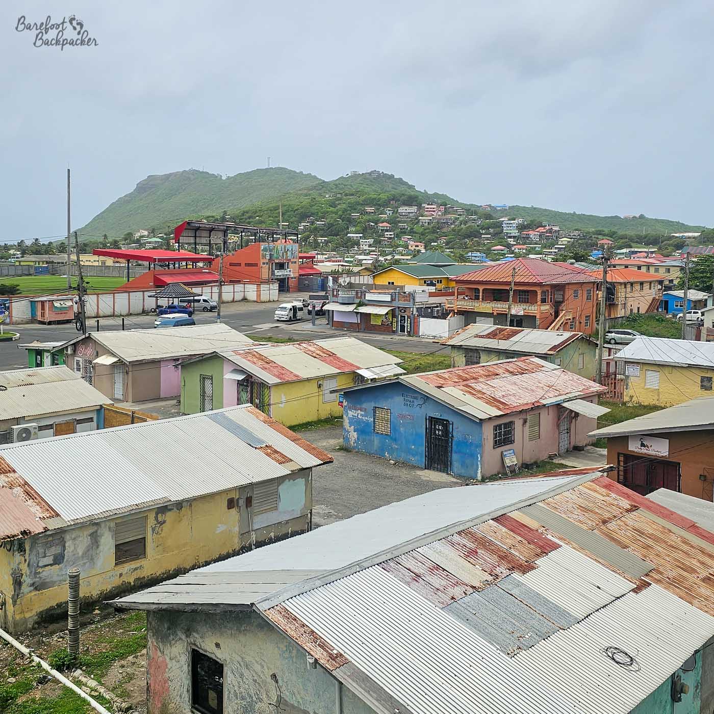 An elevated view over a residential area with small, colorful houses topped by corrugated metal roofs. In the distance, green hills rise behind larger buildings and streets under a cloudy sky.