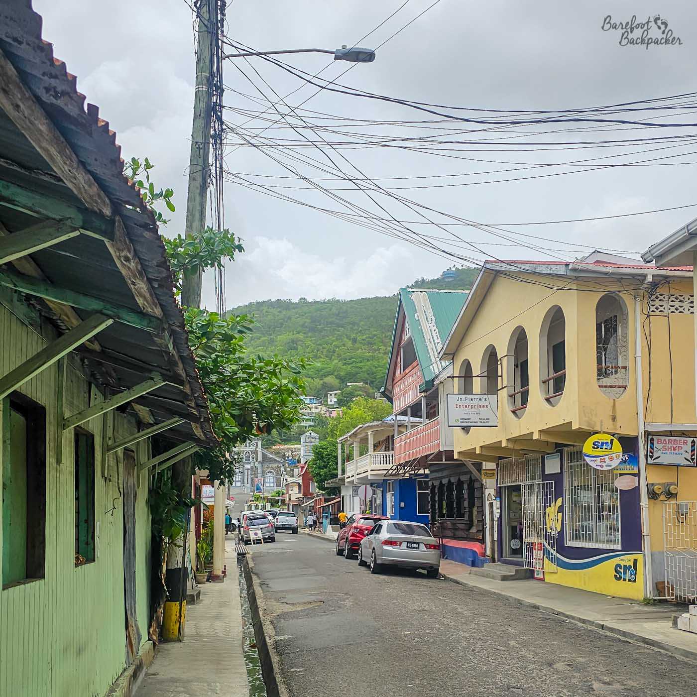 A narrow town street lined with colourful wooden and concrete buildings. Parked cars line the road, overhead power lines criss-cross above, and a green hillside rises in the background.