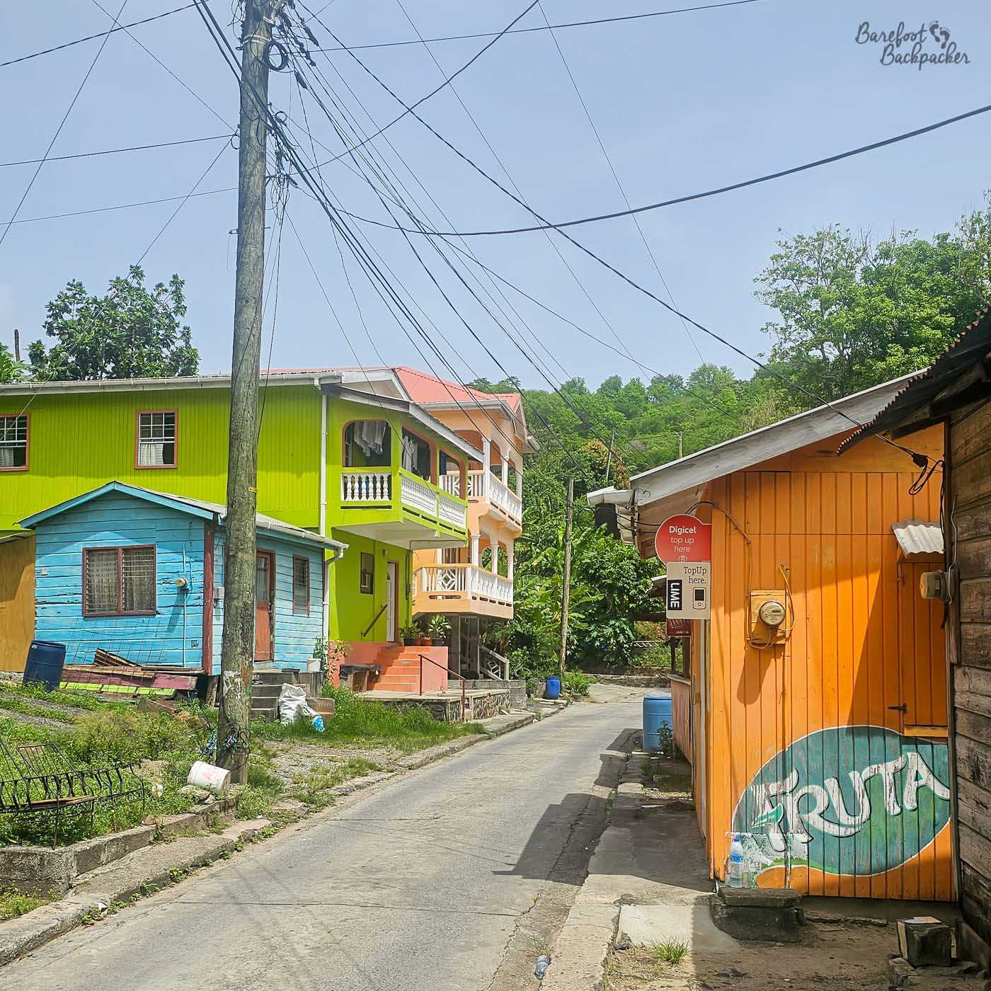 A quiet residential street with brightly painted houses in green, blue, and orange. Overhead cables stretch between poles, and dense greenery fills the background beyond the buildings.