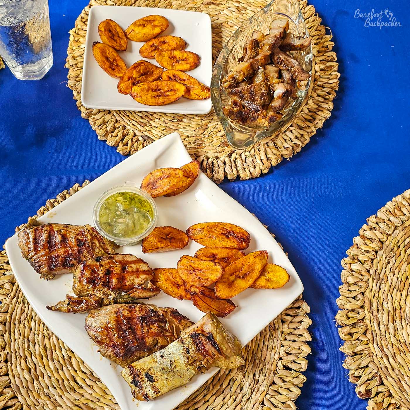 A table set with grilled meat and fried plantain on white plates. A small container of green sauce sits alongside the food, with woven placemats and a bright blue tablecloth underneath.
