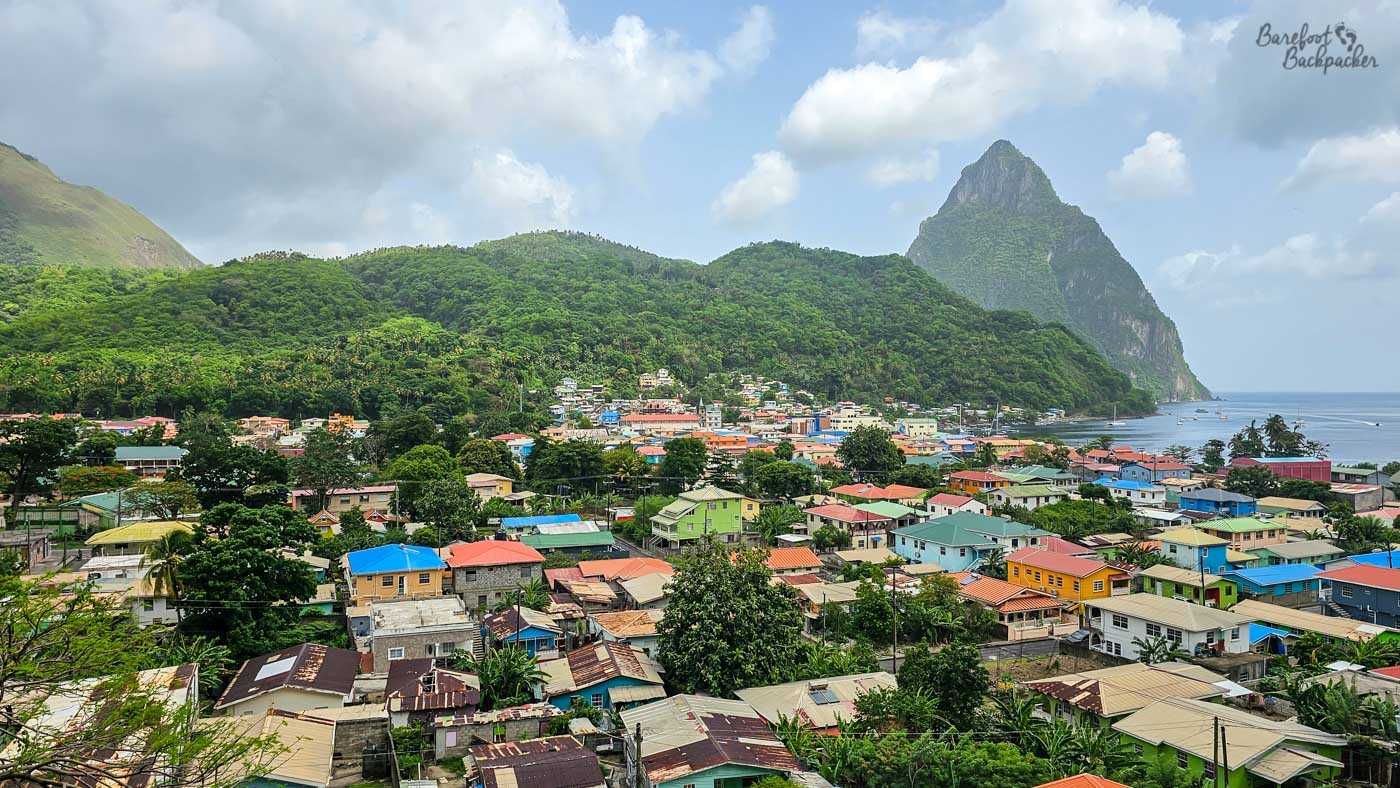 A panoramic view over a coastal town with brightly colored houses clustered below lush green hills. A steep, pointed mountain rises dramatically near the shoreline under scattered clouds.