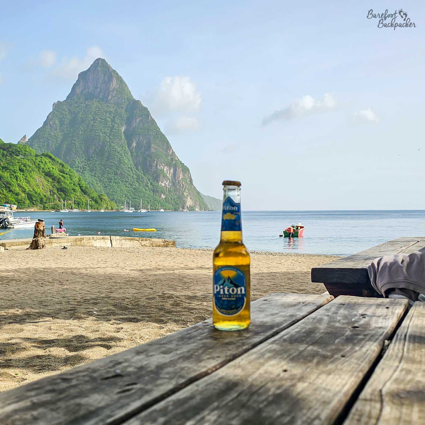A cold beer bottle standing on a wooden picnic table on a sandy beach. In the background, calm water, small boats, and a dramatic steep green mountain rise under a pale blue sky.