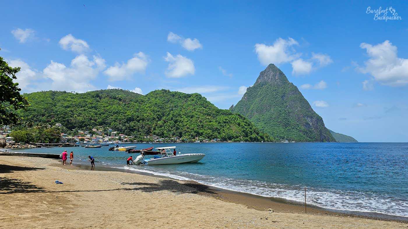 A sandy beach beside calm blue water, where small motorboats are pulled up near the shore. A steep, forested mountain dominates the background beneath a bright blue sky with light clouds.
