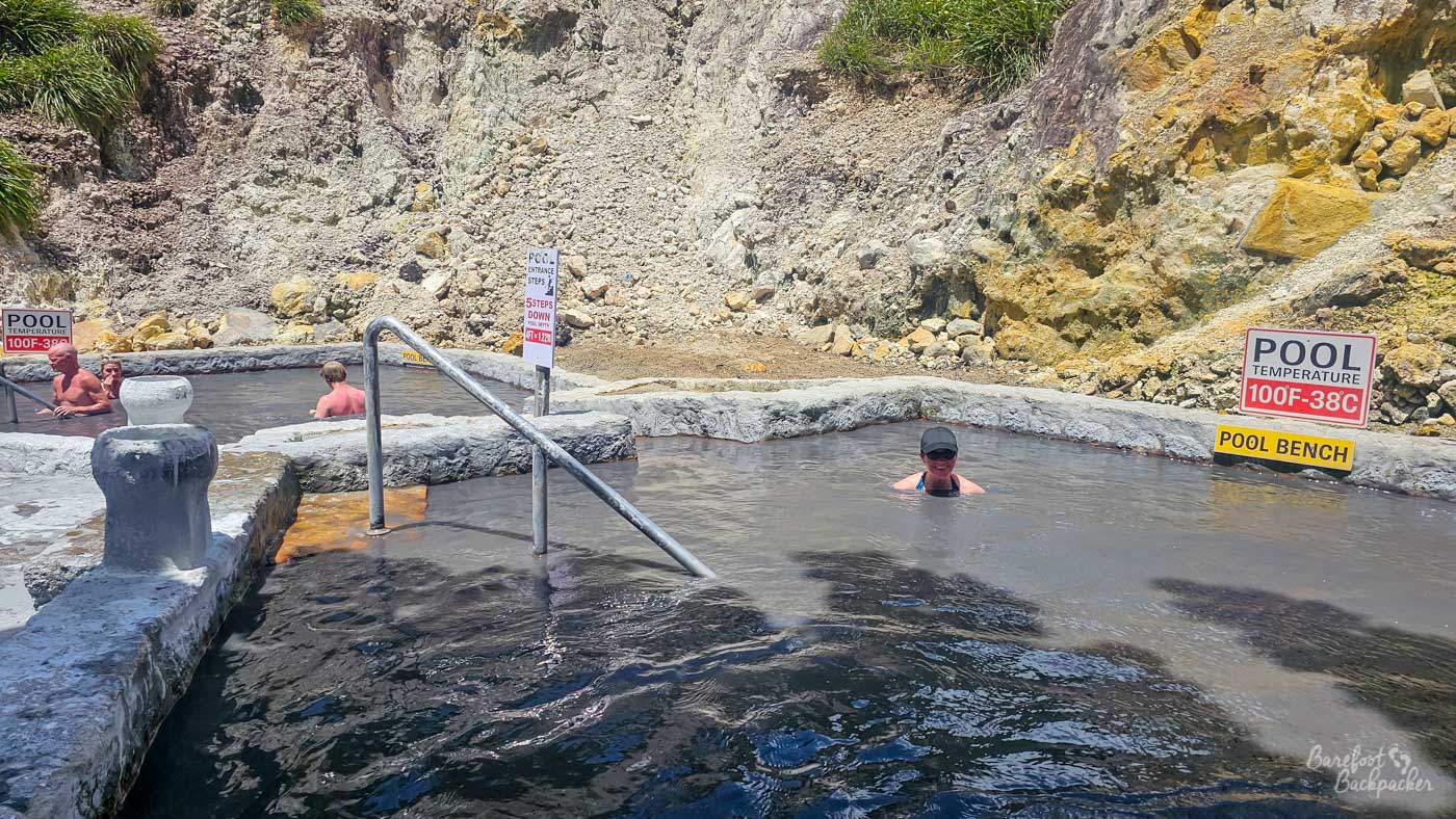An outdoor hot spring pool carved into rock, filled with cloudy mineral-rich water. People soak in the pool, and signs indicate a pool temperature of 100°F (38°C).