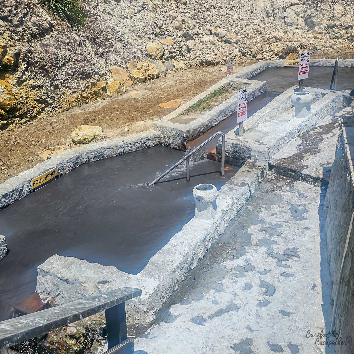 A series of stone-lined hot spring pools with steps and metal handrails, set against a rocky hillside. Signs mark pool entrances and designate pool benches.