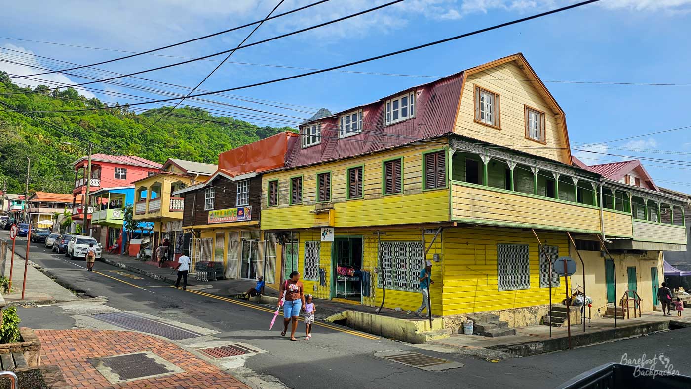 A colorful street scene with a large yellow wooden building featuring shuttered windows and an upper veranda. People walk along the road, and pastel-painted houses line the street with green hills rising behind.