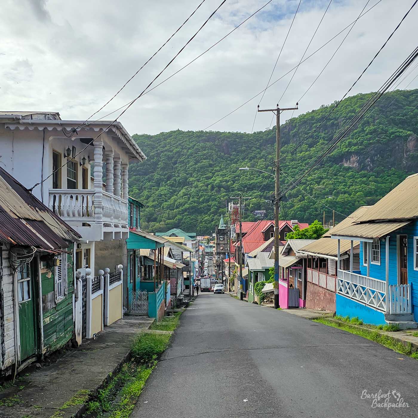 A narrow residential street lined with wooden houses painted in green, blue, pink, and white. Power lines crisscross overhead, and forested hills frame the view at the far end of the road.