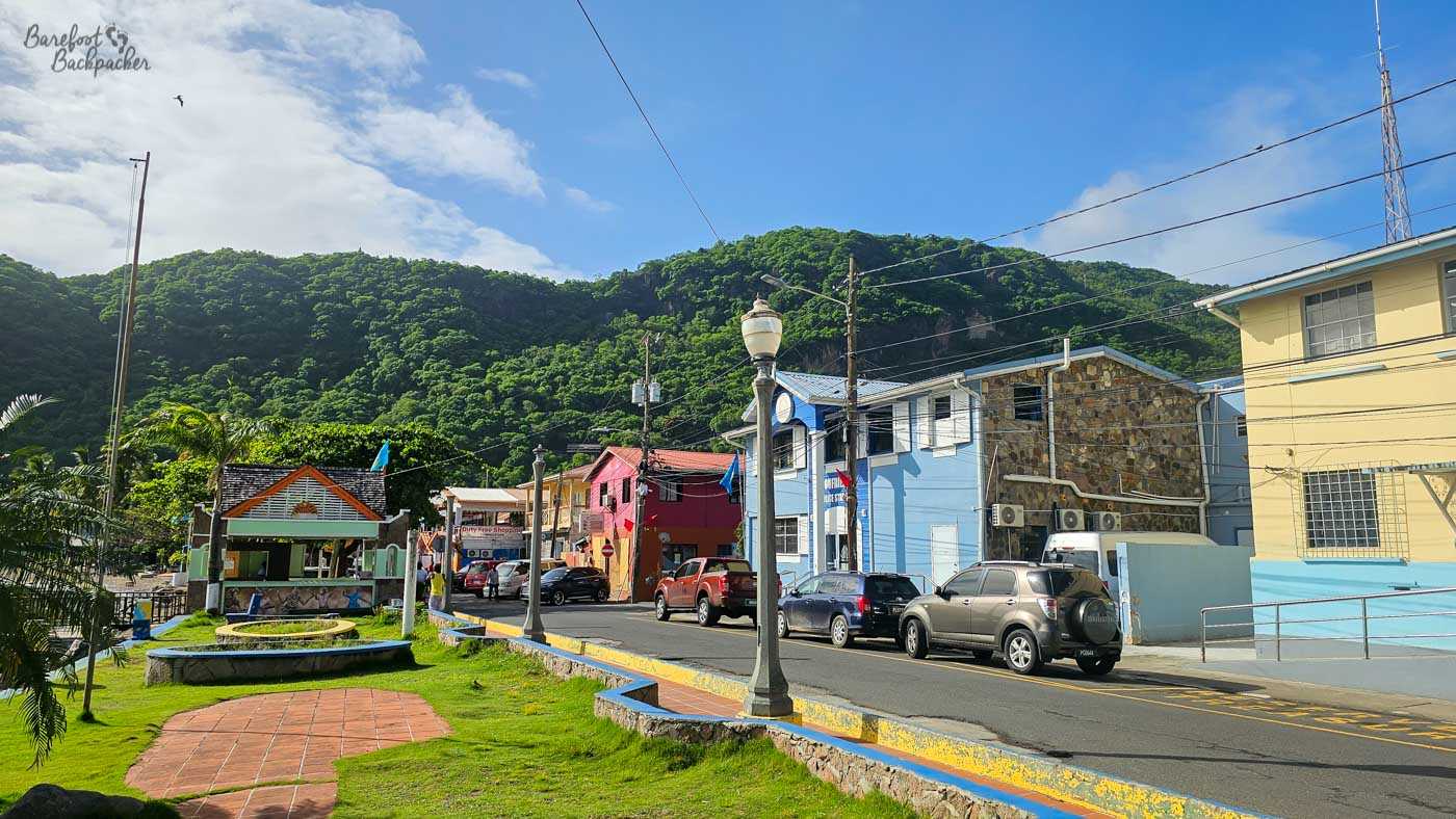 A small town square with trimmed grass, paved walkways, and a gazebo-like structure. Colorful buildings and parked cars line the street, with steep green hills forming a dramatic backdrop.