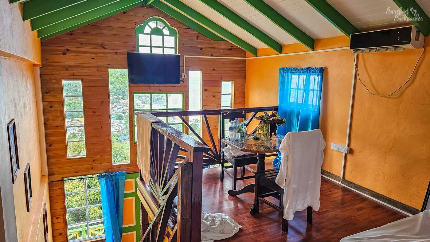 An airy loft-style interior with wooden walls, green ceiling beams, and tall windows overlooking a hillside town. A small dining table sits beside a wooden railing, with towels draped over chairs and natural light filling the space.