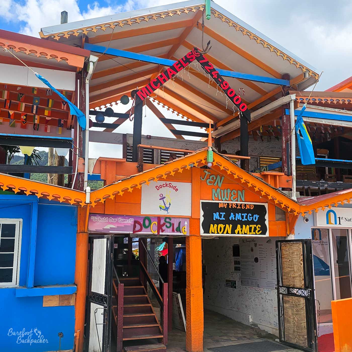 The colorful exterior of a multi-level waterfront building painted orange and blue, with signs reading “Michael’s 24,900,” “Dockside,” and multilingual greetings. A staircase leads up through the open structure beneath decorative eaves.