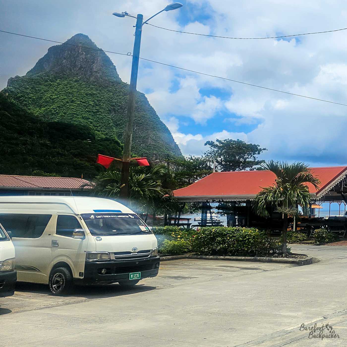 Parked minibuses and taxis sit near a waterfront area with red-roofed buildings and palm trees. A steep, forested mountain towers behind the scene beneath scattered clouds.