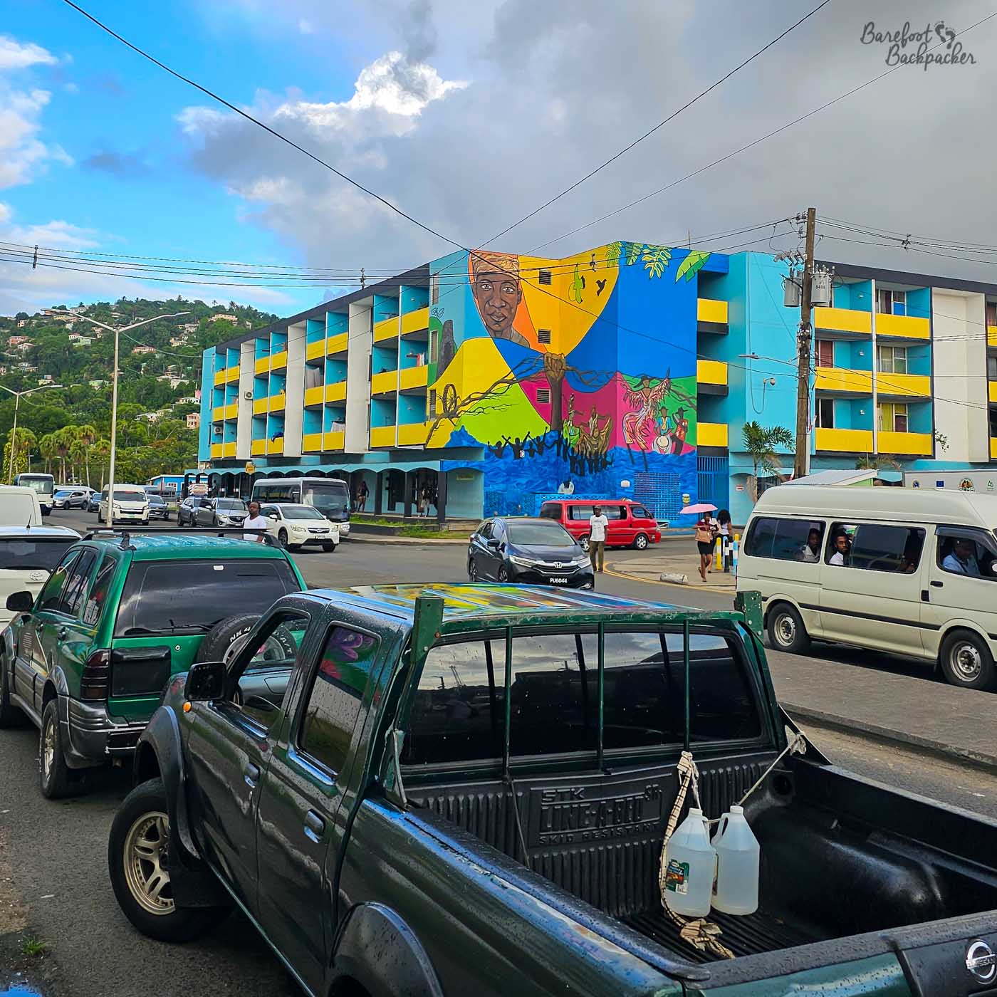 A busy street scene with cars and vans in the foreground and a large, brightly painted building covered in a colourful mural. Green hills rise behind the town under mixed blue sky and clouds.
