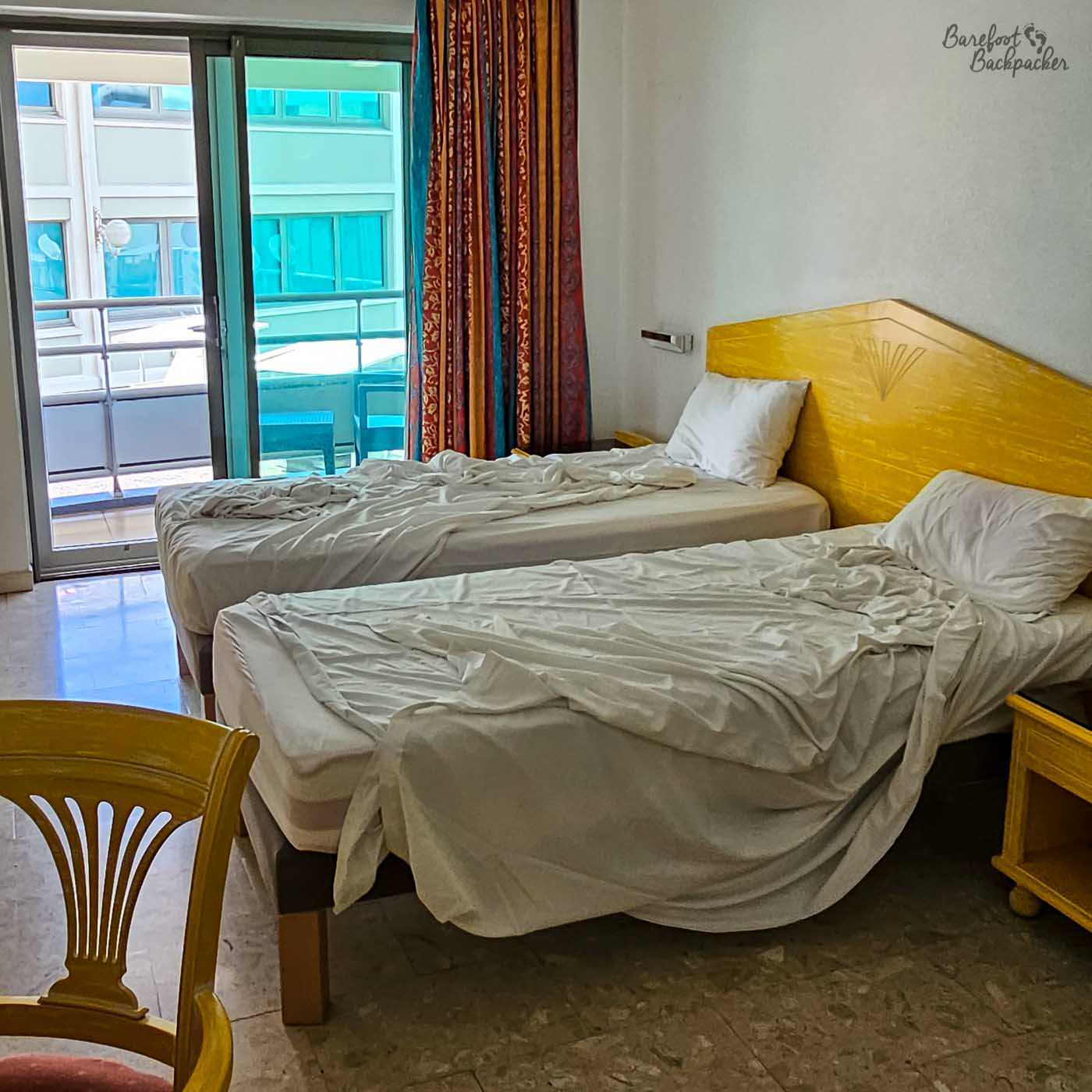 A simple hotel room with two single beds pushed together, rumpled white sheets, and wooden headboards. A sliding glass door opens onto a small balcony, letting in daylight through patterned curtains.