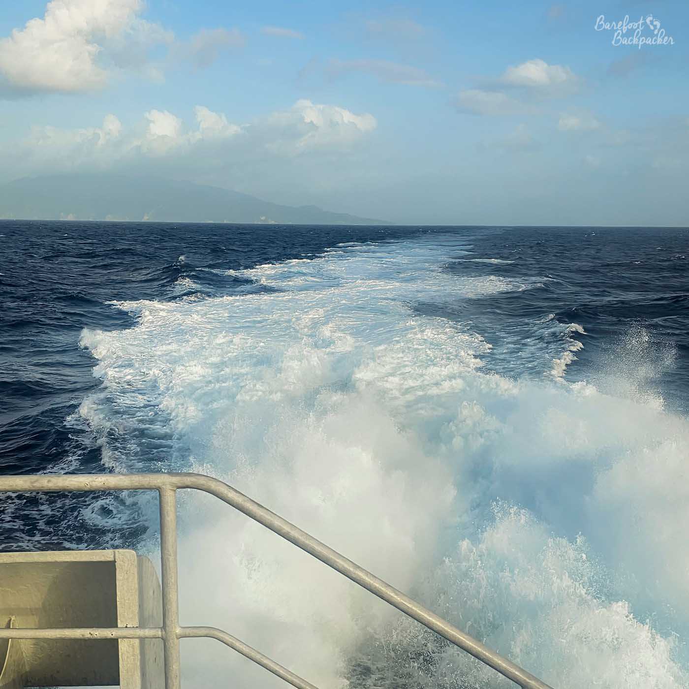 The foaming wake of a boat stretches into the distance across deep blue ocean. White spray churns in the foreground, with hazy mountains visible on the horizon beneath scattered clouds.