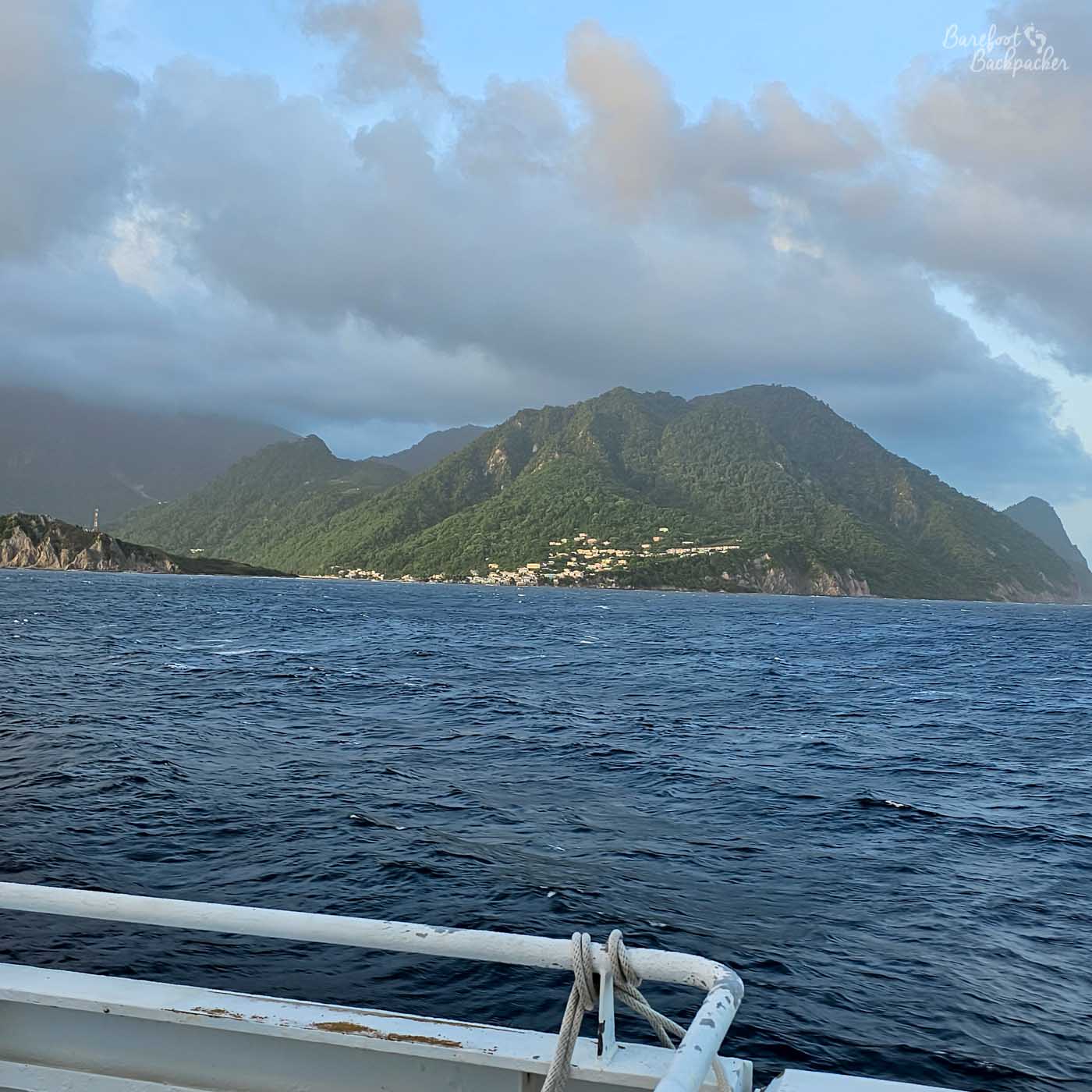 A view from a boat across deep blue ocean toward a steep, forested island coastline. Small clusters of houses dot the shoreline beneath layered grey and white clouds.