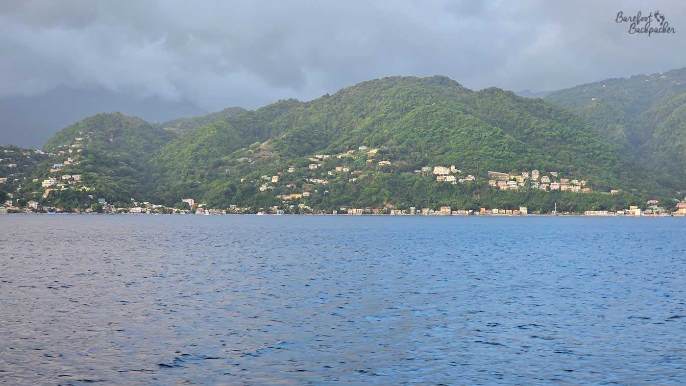 A wide coastal view across calm blue water toward a hillside town. Houses dot the green slopes beneath heavy grey clouds gathering over the mountains.
