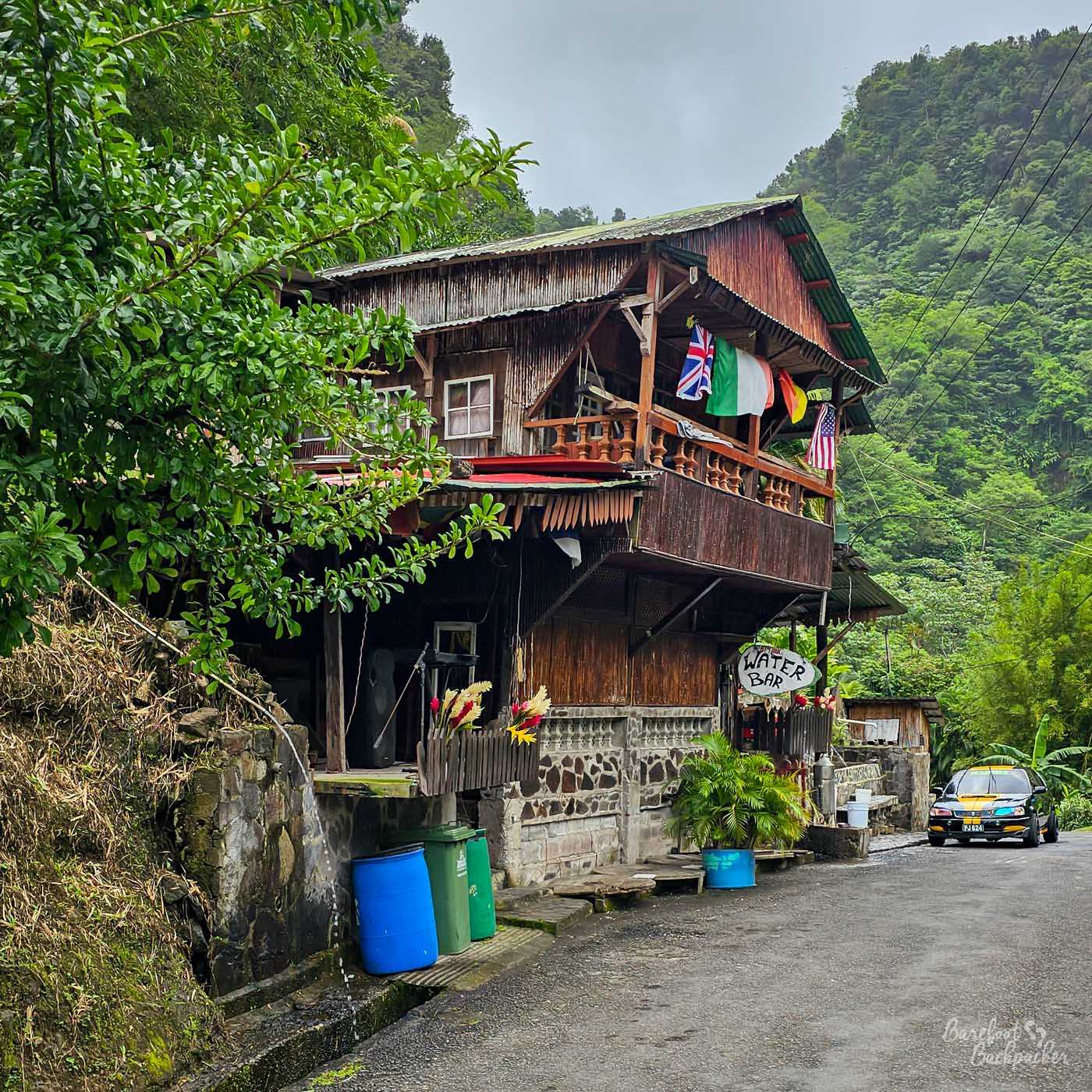 A rustic wooden building set beside a narrow road in dense rainforest. Flags hang from the upper balcony, and a hand-painted sign reads “Water Bar,” with lush green hills rising behind.