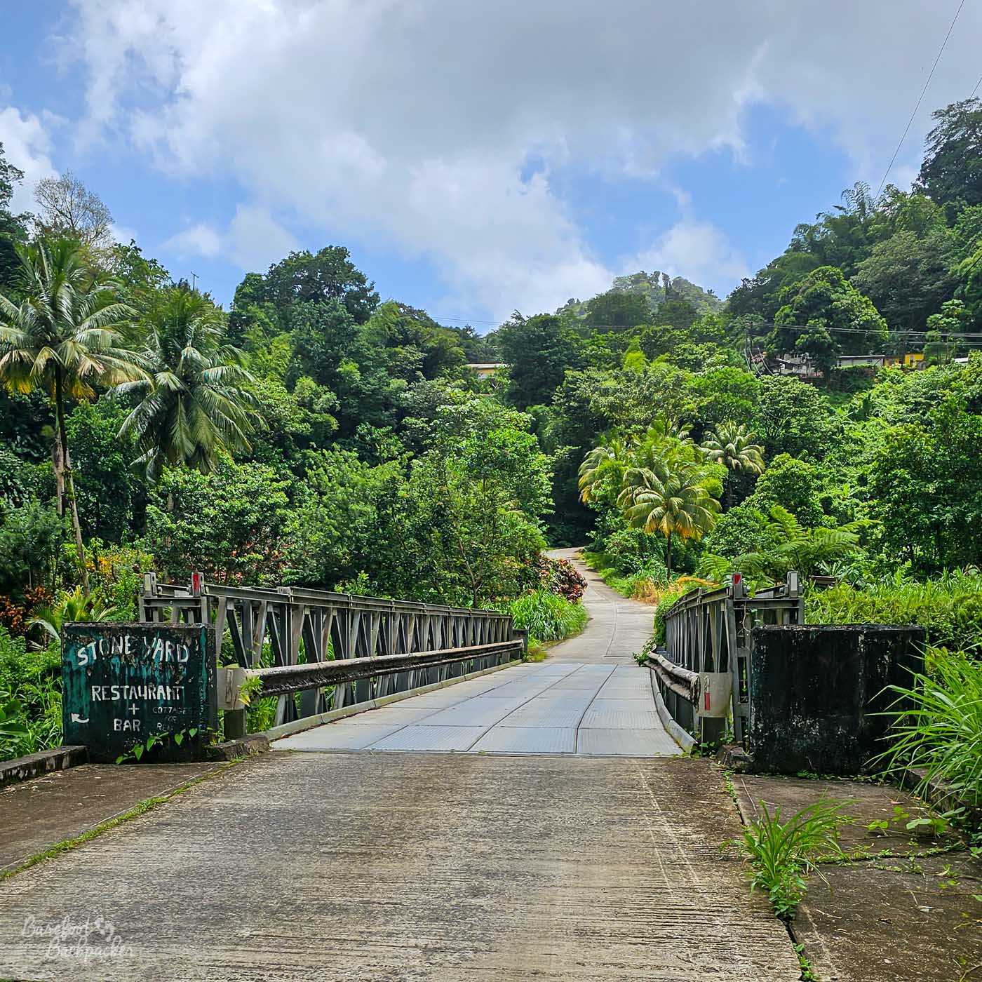 A narrow metal bridge crossing a river and leading into thick tropical forest. A sign at the entrance reads “Stone Yard Restaurant & Bar,” with dense greenery surrounding the road.