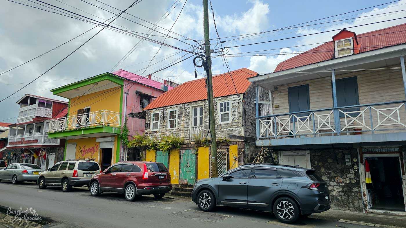 A row of older wooden buildings with balconies and peeling paint in pastel shades. Parked cars line the street, and power cables crisscross overhead.