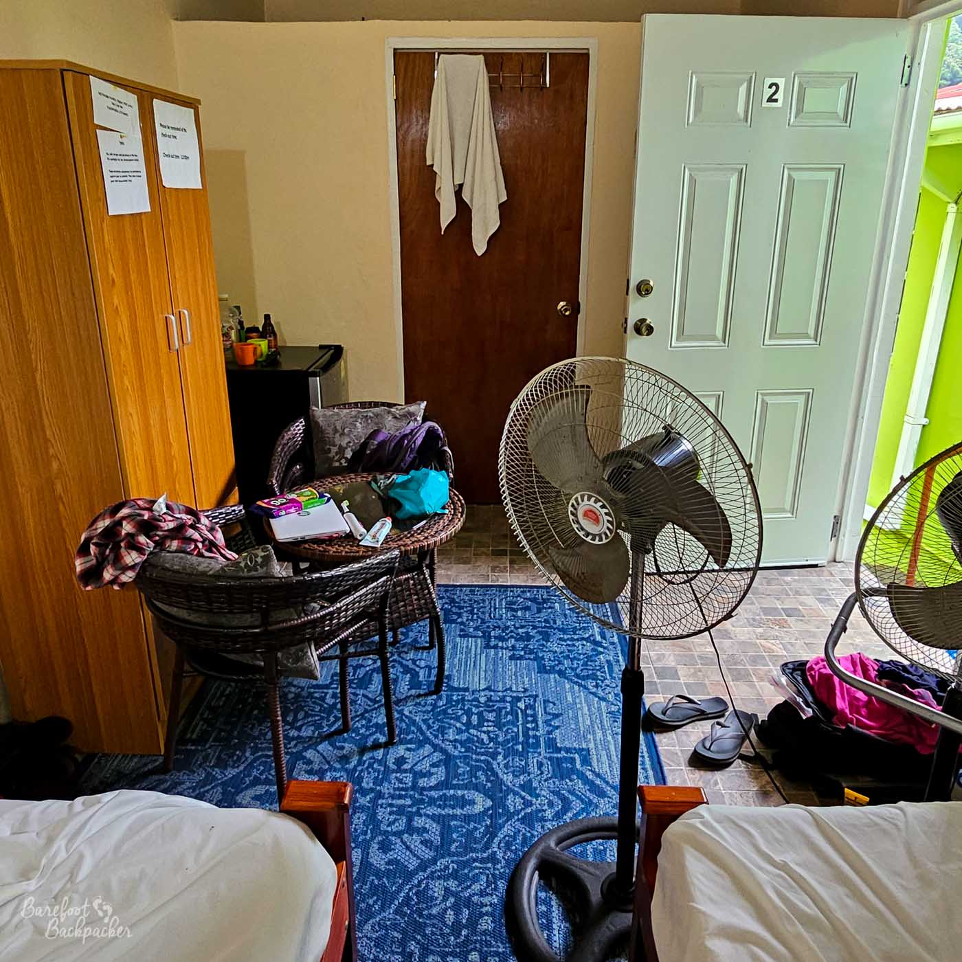 A simple guesthouse room with twin beds, standing fans, and a small table cluttered with personal items. An open door lets daylight into the tiled room.