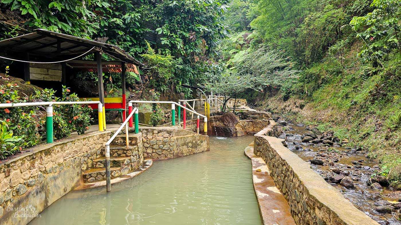A series of stone-lined hot spring pools filled with cloudy mineral water. Narrow walkways with handrails wind through dense tropical vegetation surrounding the pools.
