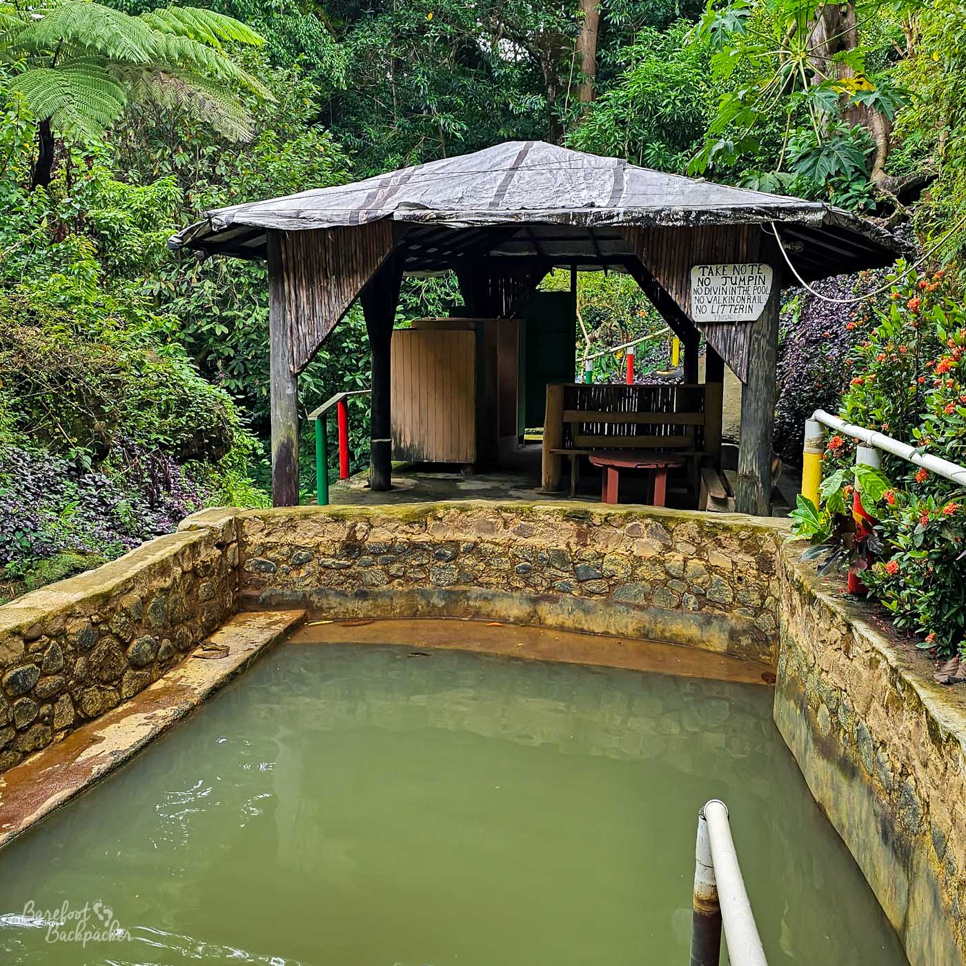 A circular stone hot spring pool beneath a simple wooden shelter. A sign lists rules for pool use, and lush rainforest plants surround the structure.