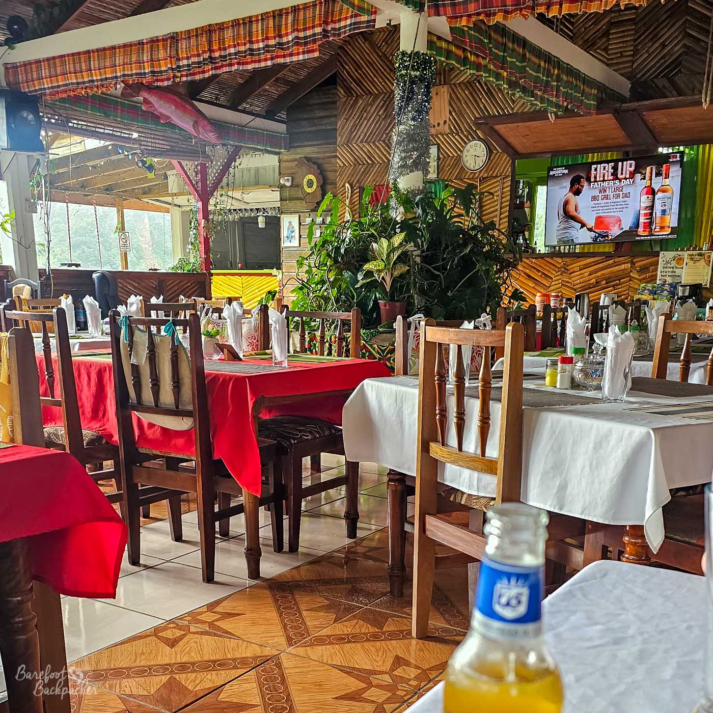 An open-air restaurant interior with wooden furniture and red-and-white tablecloths. Potted plants hang and cluster around the dining area, and a television plays above the bar in a warm, tropical setting.
