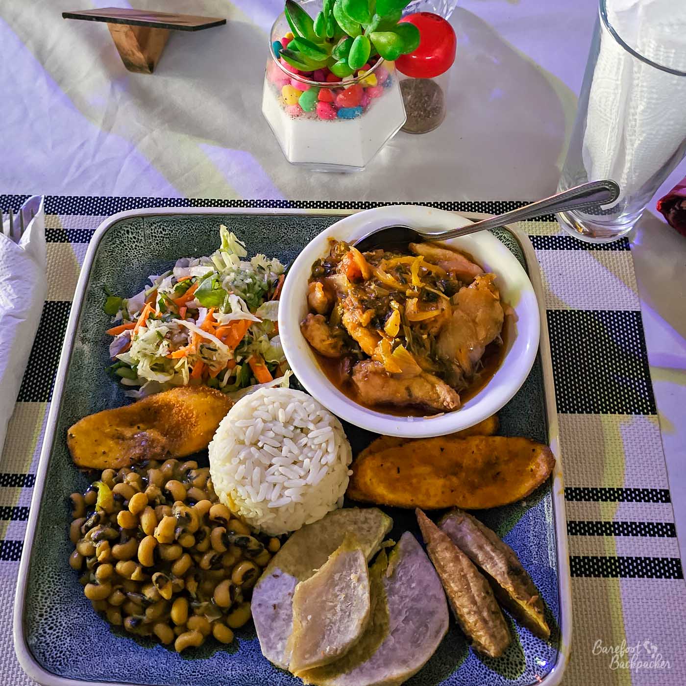 A large square plate of Caribbean food featuring stewed chicken in sauce, white rice, fried plantain, black-eyed peas, coleslaw, and boiled ground provisions. The plate sits on a woven placemat beside condiments.