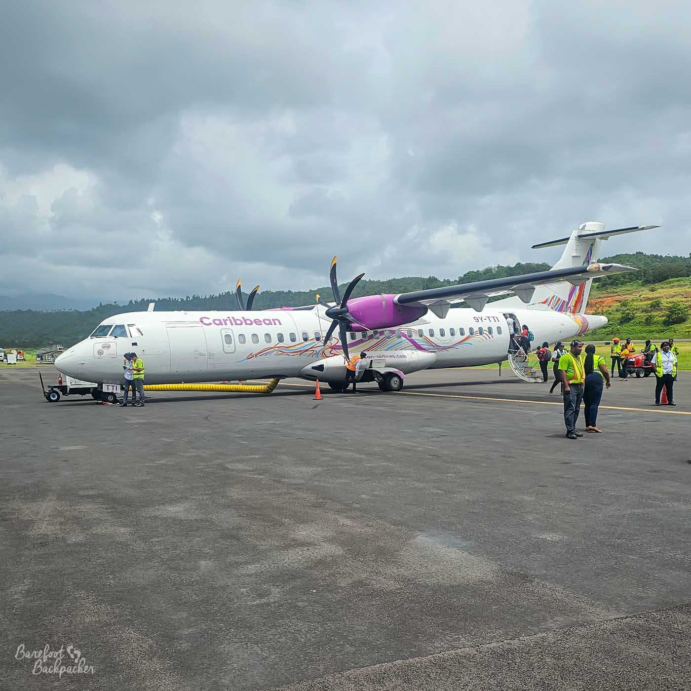 A Caribbean Airlines turboprop aircraft parked on a small airport runway. Ground crew in high-visibility vests stand nearby as passengers board via a rear stairway under an overcast sky.