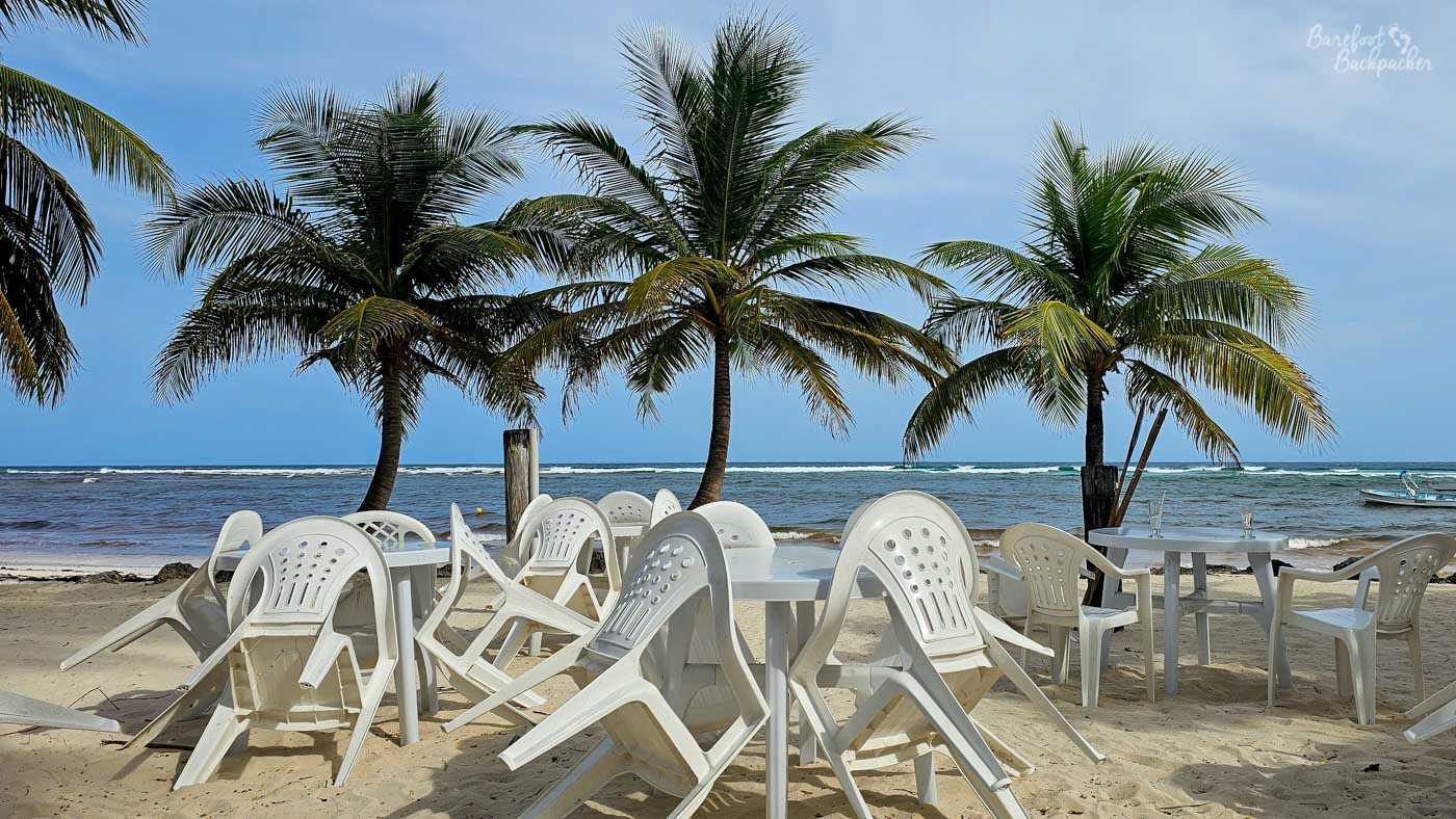 White plastic tables and chairs scattered on a sandy beach beneath tall palm trees. The sea lies just beyond, with small waves rolling in under a pale blue sky.