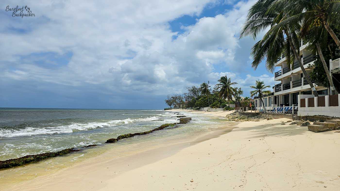 A quiet sandy beach lined with palm trees and low seaside buildings. Gentle waves wash over rocky outcrops along the shoreline, and the sky is partly cloudy with soft light.