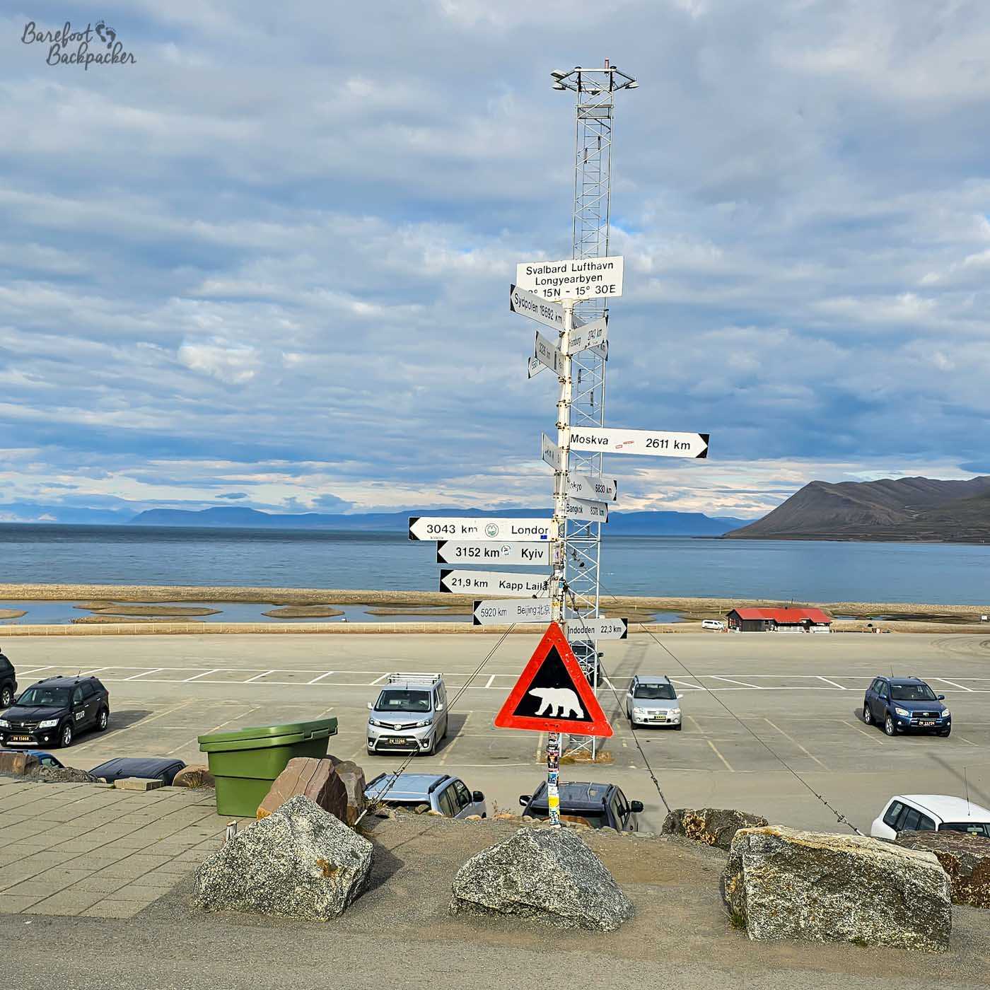 A tall signpost outside Longyearbyen Airport points to cities around the world with distances, including Moscow, London, and Kyiv. A red triangular polar bear warning sign stands in front, with parked cars, the runway, and a calm fjord backed by mountains beyond.
