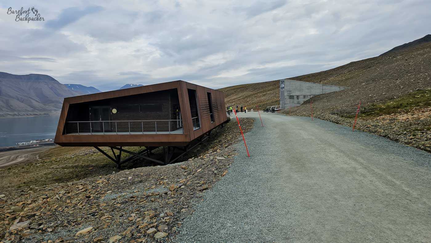 A modern, angular building clad in weathered steel juts dramatically out over a rocky slope, supported by dark metal stilts that lift it above the ground. Its sharp, geometric form contrasts with the soft curves of the surrounding mountains and the calm, silvery water of a fjord visible below. A gravel road winds past the structure, marked by thin red poles, where a small group of people walks toward a concrete entrance set into the hillside. The entire scene feels stark and windswept, blending contemporary design with the raw, rugged beauty of the Arctic environment.