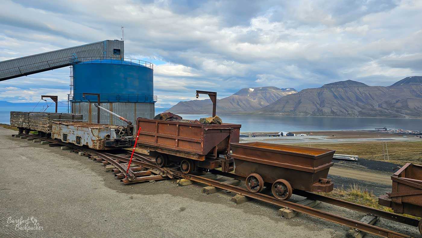 Rusting mining carts sit on narrow-gauge tracks beside a large blue storage tank, remnants of Svalbard’s coal mining past. The scene looks out over the airport, fjord, and layered mountains under a cloudy sky.
