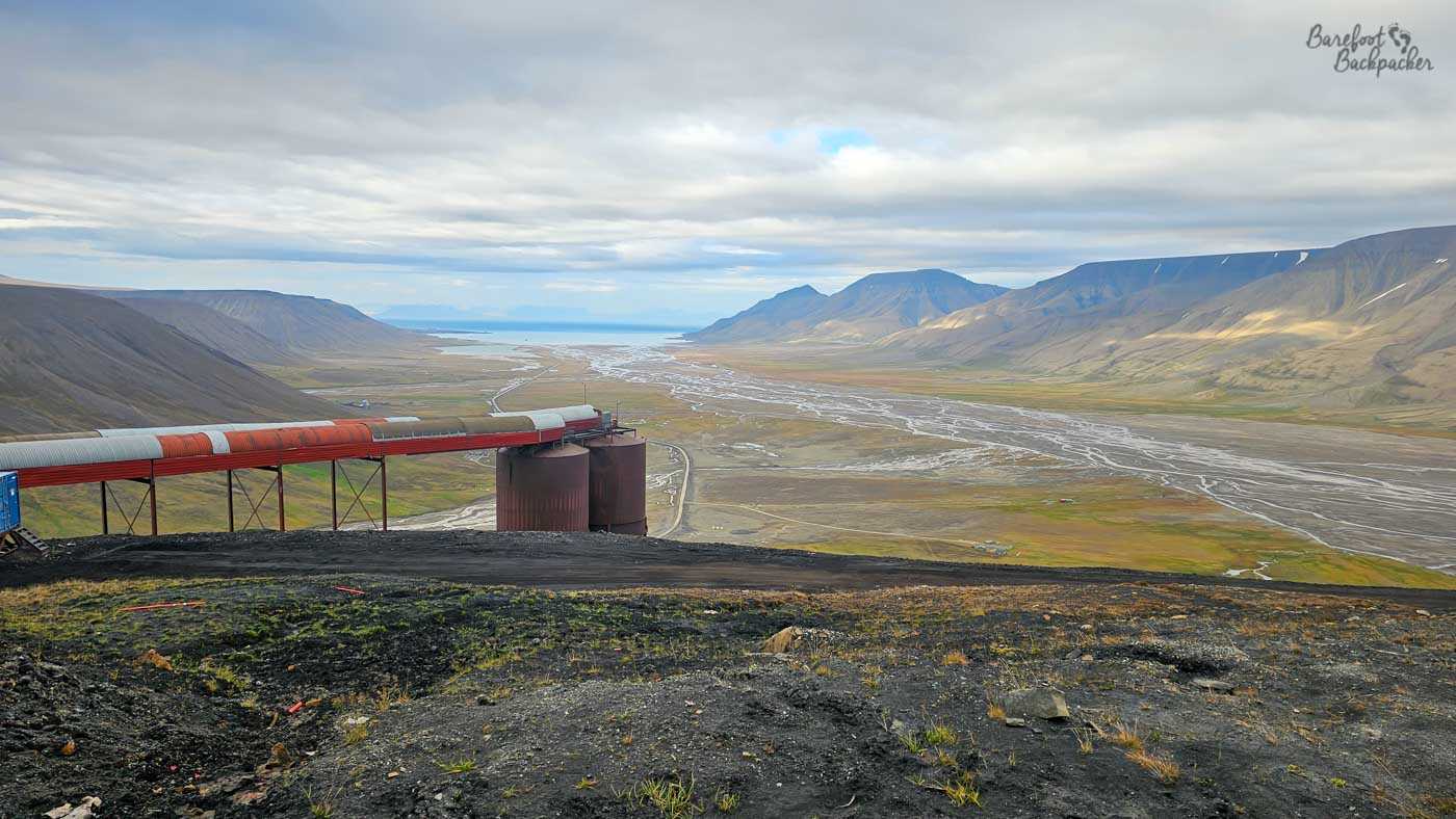 From a high vantage point, a vast valley opens out toward a distant strip of water, its floor crisscrossed by winding, braided river channels that glint faintly under diffuse light. In the foreground, a rust-red industrial conveyor or pipeline stretches across the slope, supported by metal frames and leading toward large cylindrical tanks. The surrounding mountains rise steeply on either side, their muted tones of grey, brown, and green softened by low cloud cover. The scale of the landscape dwarfs the industrial elements, emphasising both the harshness of the environment and the effort required to operate within it.