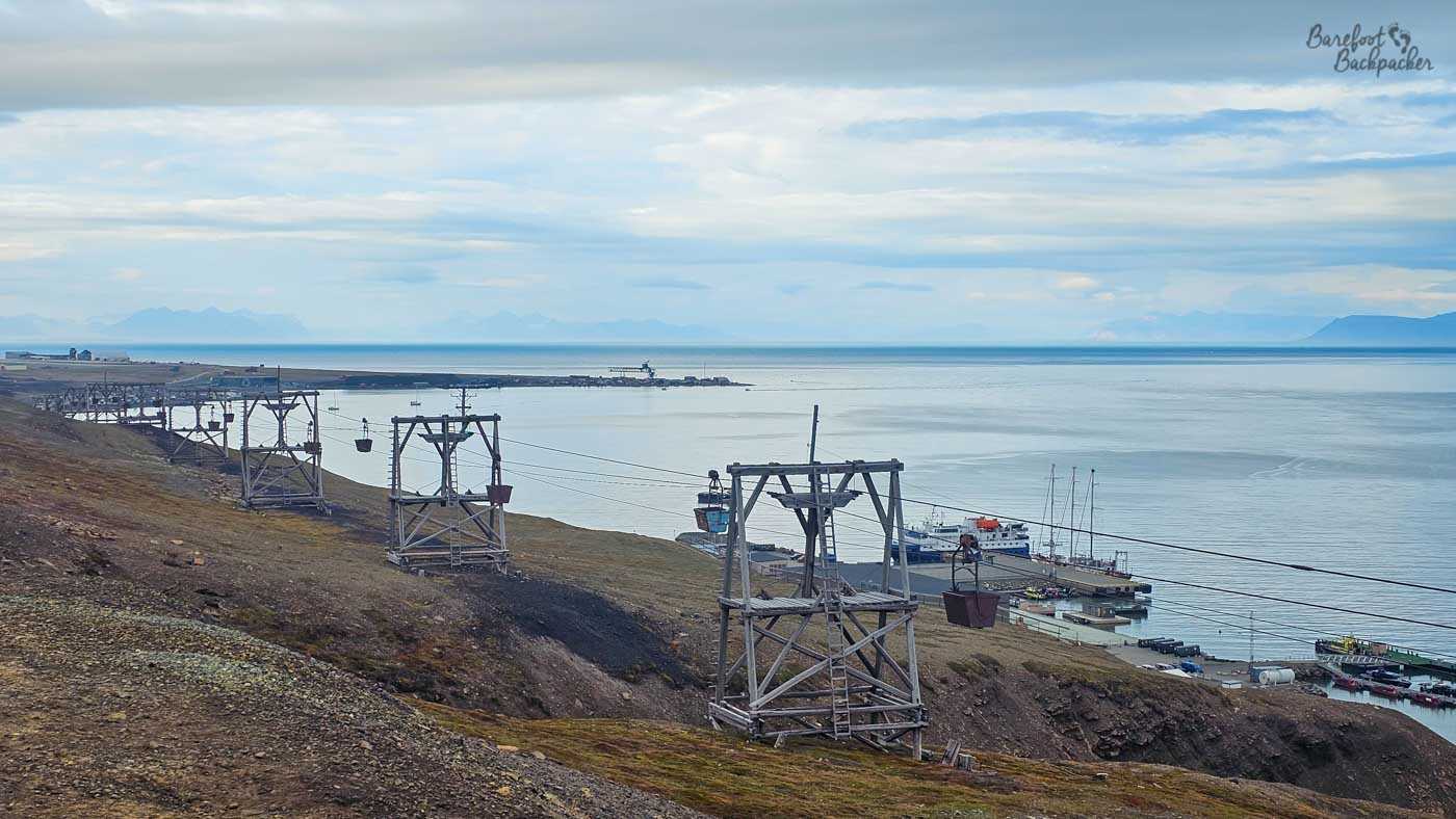 A line of weathered wooden cableway towers marches down a barren hillside toward the sea, their hanging buckets once used to transport coal now suspended over a quiet Arctic harbour. Below, a small port with moored boats juts into calm, steel-grey water, with distant mountains fading into the horizon under a heavy sky.