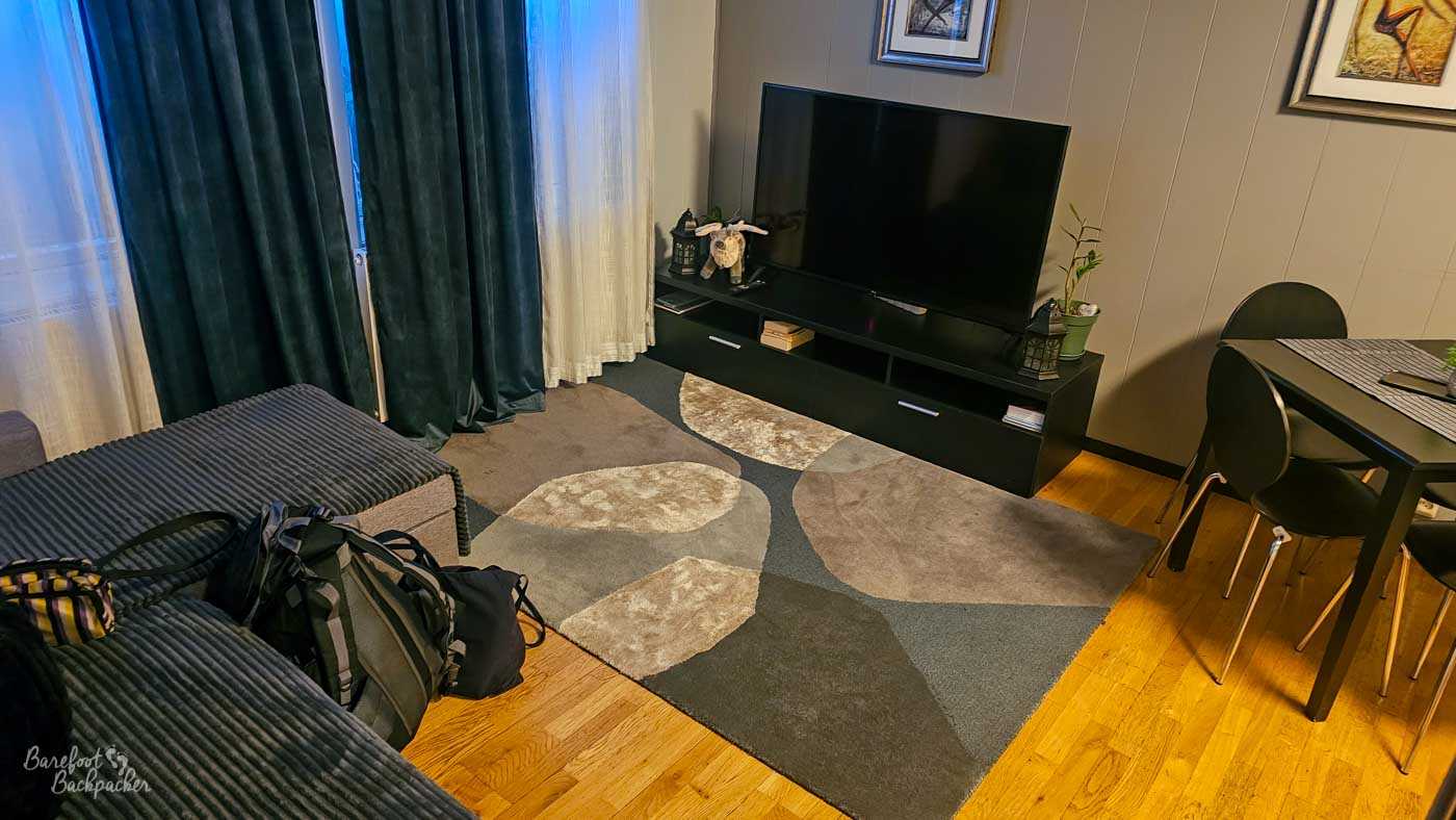 A small, tidy living space with a sofa, patterned rug, and flat-screen TV on a low unit. A dining table sits to the side, and a backpack rests on the sofa, suggesting a traveller settling in.