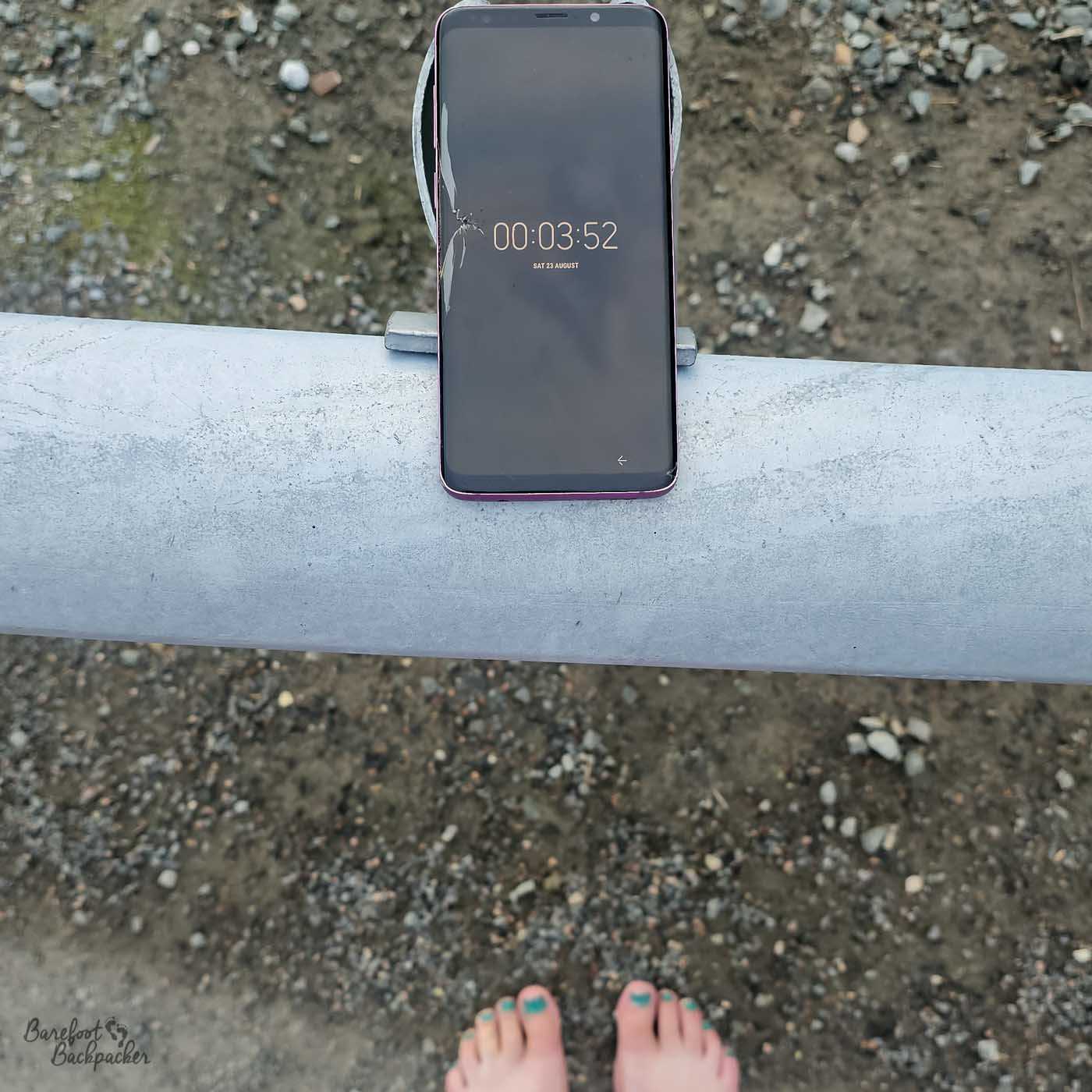 A smartphone balanced on a railing displays the time just after midnight, while daylight still illuminates the rocky ground below. Bare feet visible at the bottom of the frame emphasise the surreal midnight sun conditions.