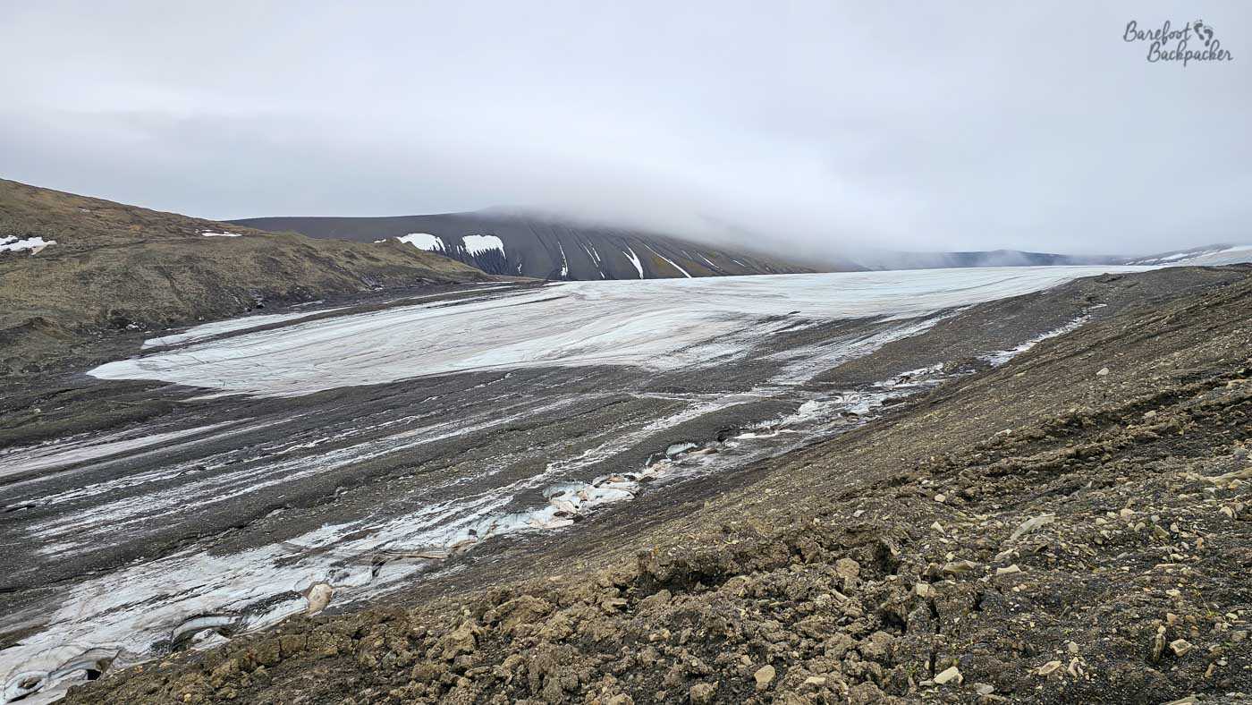 A broad, gently sloping glacier stretches across the frame, its pale, streaked surface cutting through a barren valley of dark rock and gravel. The ice is marked with lines of debris and meltwater channels, giving it a textured, almost striped appearance. Low clouds hang heavily over the surrounding hills, partially obscuring the higher ridges and softening the horizon. The overall scene feels cold and subdued, with the glacier quietly dominating an otherwise stark, lifeless landscape.
