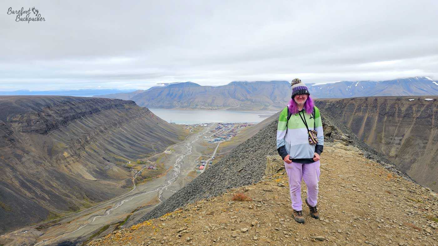 A lone figure stands on a narrow, rocky ridge high above a wide Arctic valley, with a small settlement and fjord visible far below. The person, bundled in layers with a hat and colourful clothing, provides a vivid contrast to the muted tones of the surrounding mountains. Steep slopes drop away on either side, leading down to braided river channels that wind toward the water. The scale of the landscape is immense, making the human presence feel both adventurous and fragile against the vast terrain.