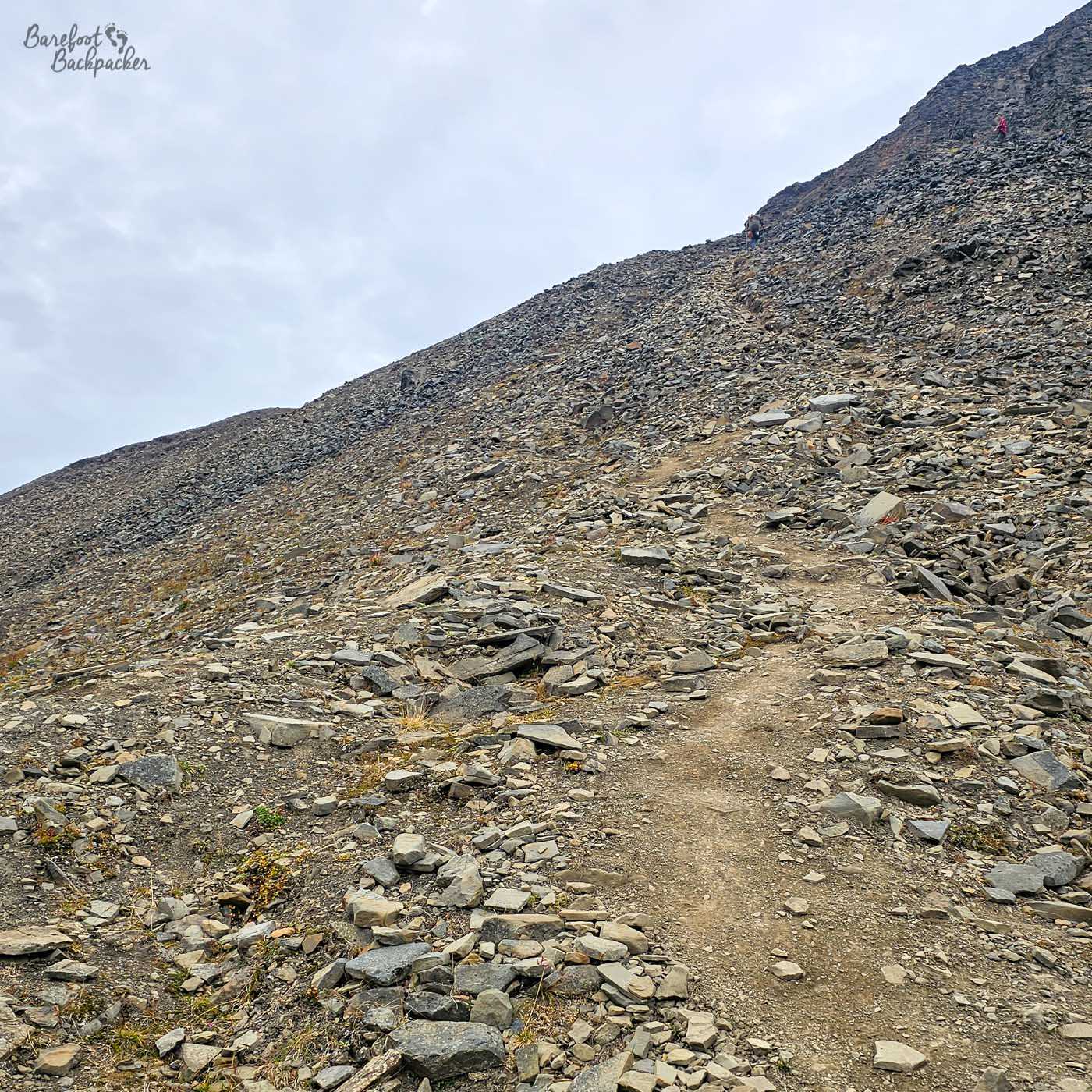 A rough hiking path winds diagonally up a steep hillside covered in loose, broken rocks of varying sizes, creating a challenging and uneven surface. The slope is almost entirely barren, with only occasional patches of hardy vegetation clinging to the ground. A few distant figures can be seen higher up, emphasising the incline and the effort required to climb it. The sky above is pale and overcast, adding to the stark, rugged feel of the terrain.
