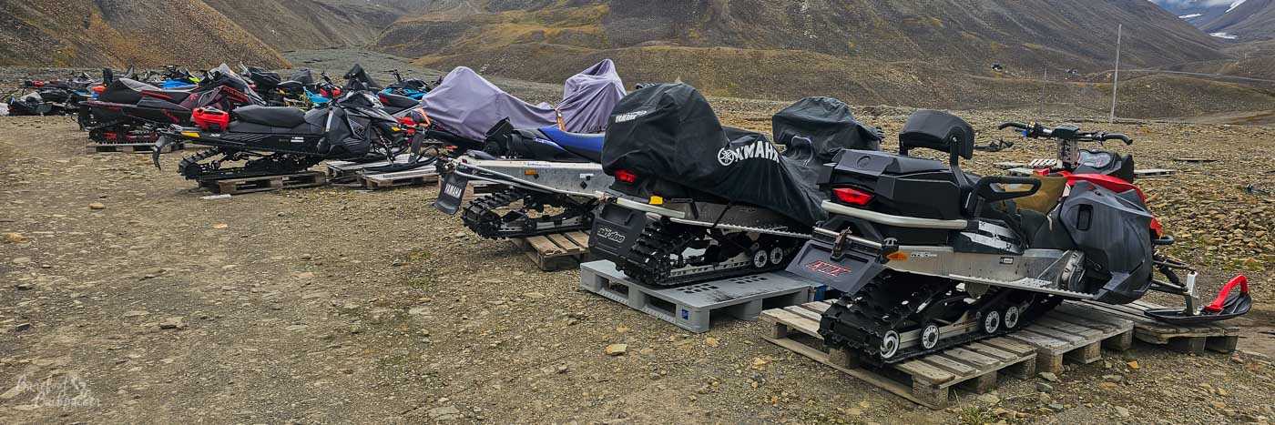 A long row of snowmobiles, some covered and some exposed, are neatly parked on wooden pallets across a gravelly Arctic plain. Their rugged tracks and gear hint at expeditions into the surrounding mountains, which rise stark and treeless under a cool, overcast sky.
