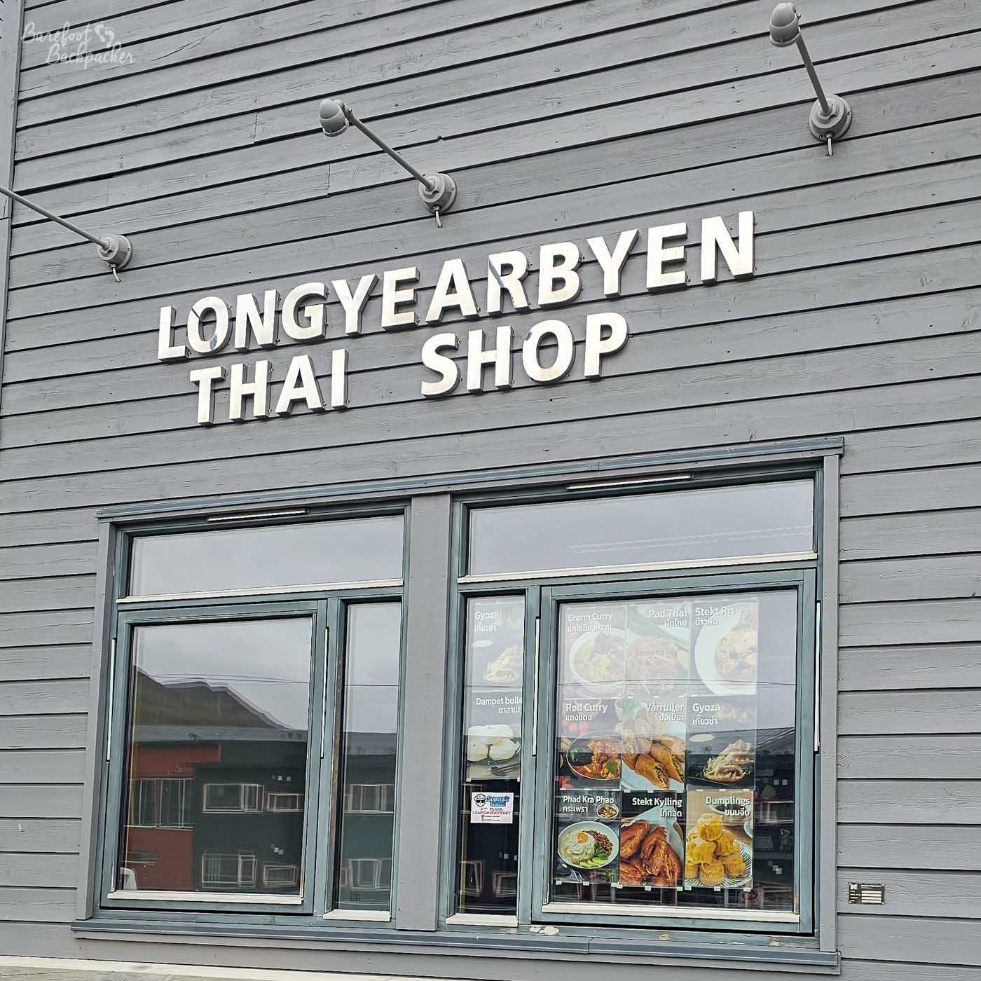 Close-up of a grey wooden building in Longyearbyen with bold white lettering reading “Longyearbyen Thai Shop.” Below the sign, large windows display a menu with photos of Thai dishes, adding a splash of colour to the otherwise muted Arctic streetscape.