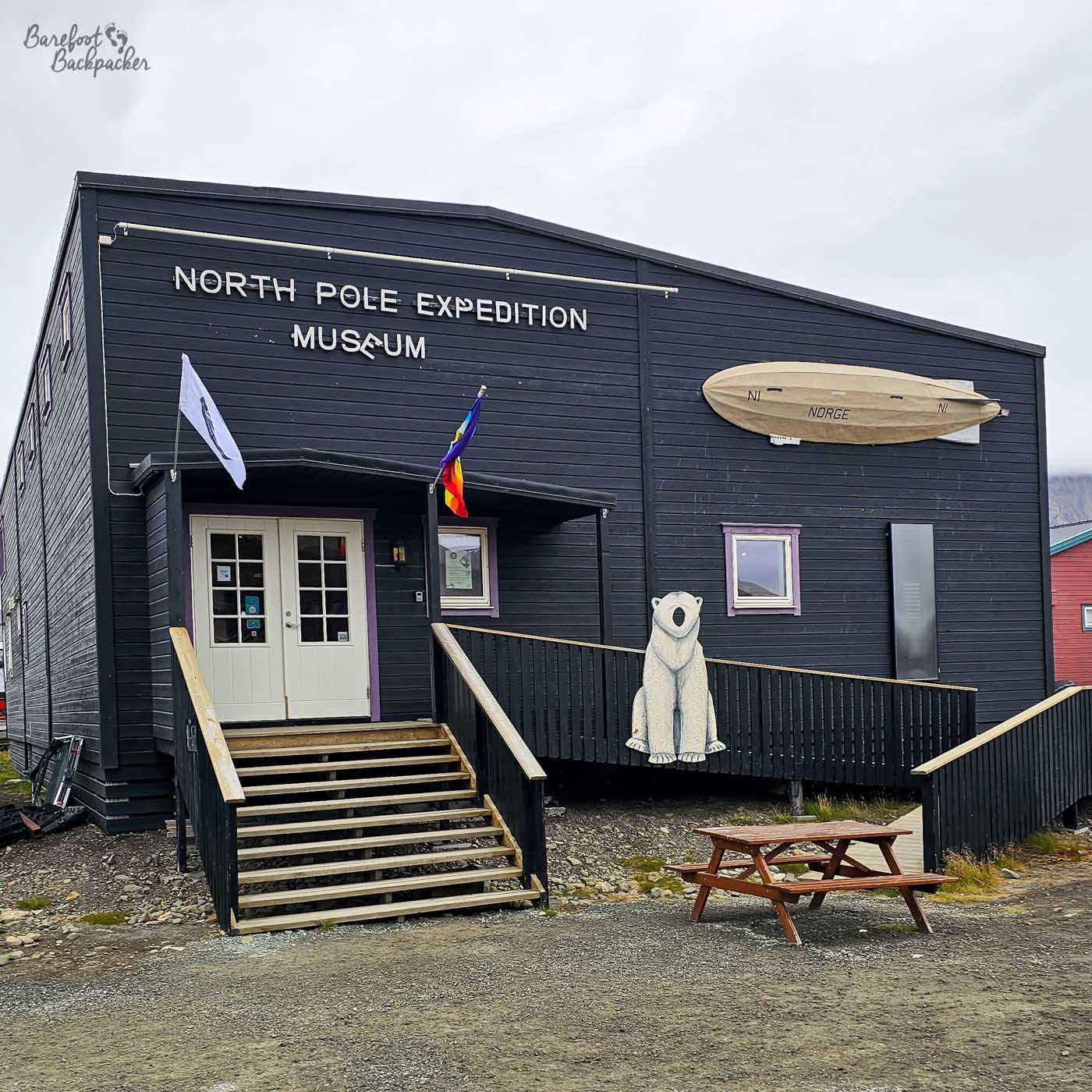 A dark wooden building labelled “North Pole Expedition Museum” stands under a grey Arctic sky, its simple facade accented by white lettering and a mounted model of an airship on the wall. A short staircase leads up to double doors, while a ramp runs alongside, where a small white polar bear statue sits as a playful detail. Flags flutter at the entrance, adding a splash of colour against the otherwise muted tones. In front, a wooden picnic table rests on the gravel, reinforcing the remote, practical feel of the setting.
