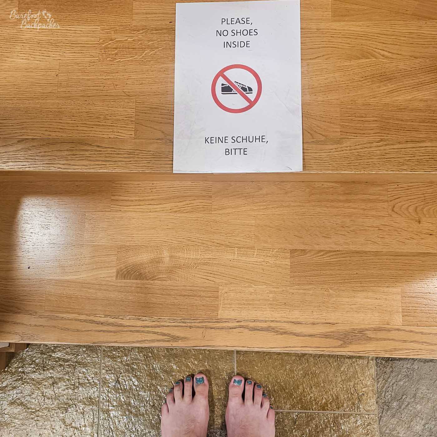 A printed sign on a wooden floor reads “Please, no shoes inside” (also in German), with a crossed-out shoe symbol. At the bottom of the frame, a pair of bare feet with painted toenails stands on the threshold, complying with the rule.