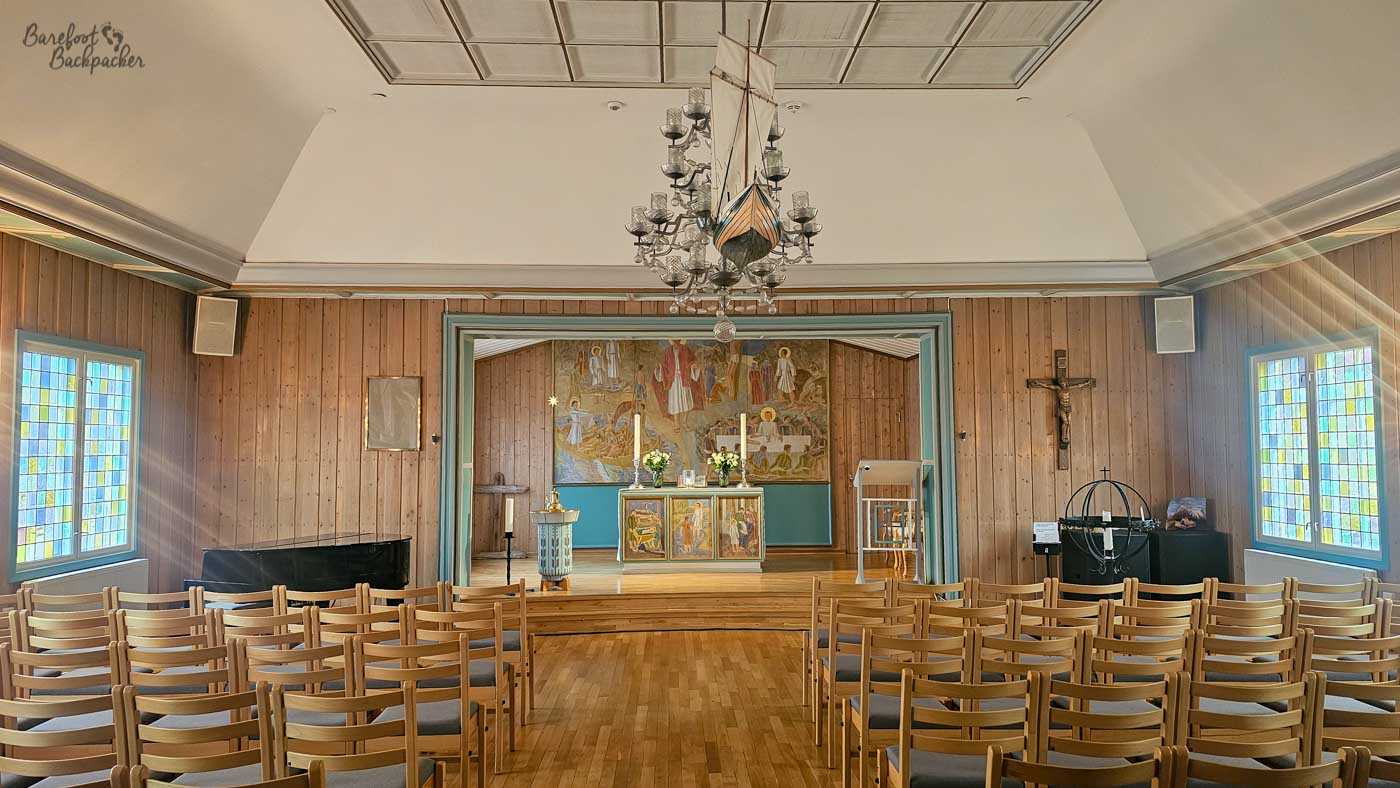 Inside a small wooden chapel, rows of simple light-coloured chairs face a modest altar framed by a painted mural depicting religious figures. Soft daylight filters through stained glass windows on either side, casting gentle colour onto the walls and floor. A chandelier hangs from the ceiling, adding a touch of elegance to the otherwise understated space. The atmosphere feels शांत and intimate, with clean lines and warm wood creating a calm, reflective environment.