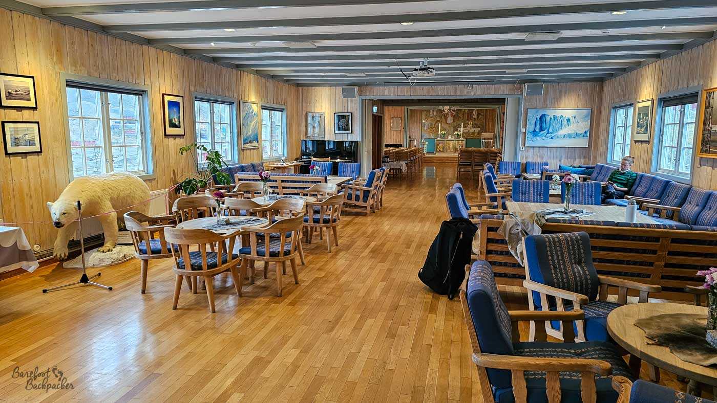 A warm, wood-panelled lounge with tables and blue-upholstered seating, leading through to a small chapel at the far end. A taxidermy polar bear stands roped off to one side, while a few people relax inside the quiet space.